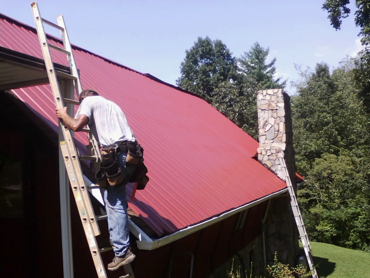 Roofer on a ladder beside a red metal roof and stone chimney — Soffit & Fascia Repair by Roofing Brothers in Helena